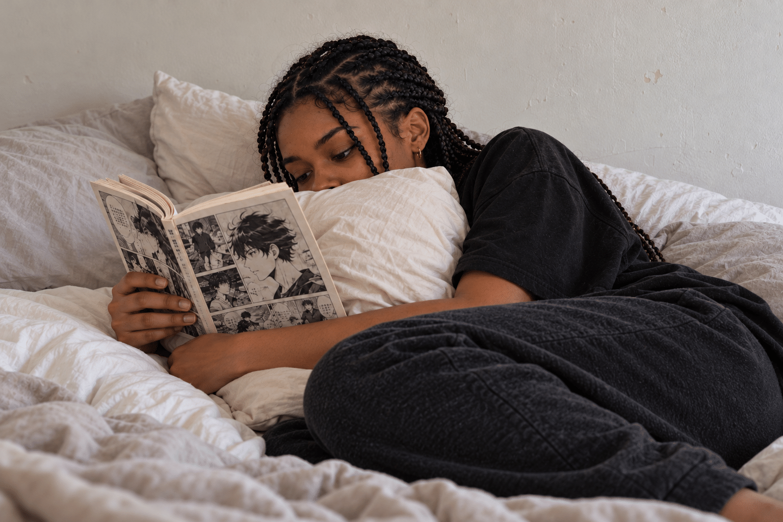 Young woman with box braids lying in bed hugging a pillow while reading a manga volume